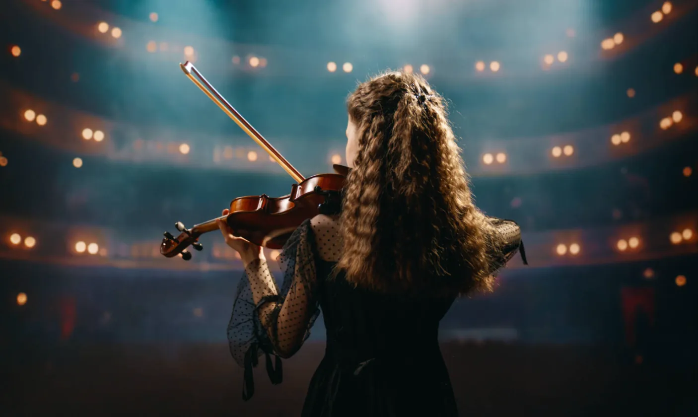A violinist seen from behind, facing a lit concert hall
